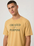 A man wearing a mustard yellow t-shirt with the printed text 'CREATED with a PURPOSE' in dark green, photographed against a neutral light gray background.