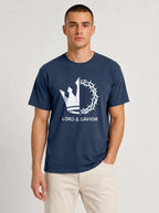 A man wearing a navy blue crewneck t-shirt with a white graphic of a crown of thorns and the text 'LORD & SAVIOR' on the front, paired with light beige pants, against a light gray studio background.