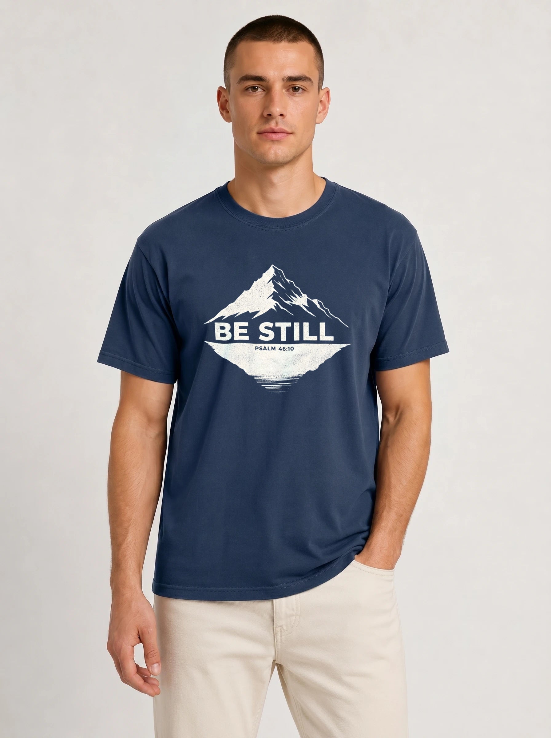 A man wearing a navy blue crewneck t-shirt with a white graphic of a mountain and the text 'BE STILL' and 'Psalm 46:10' on the chest, paired with light beige pants, against a light gray studio background.