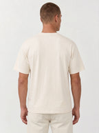 A man viewed from the back wearing a plain cream-colored short-sleeve t-shirt and matching cream pants against a light gray studio background.