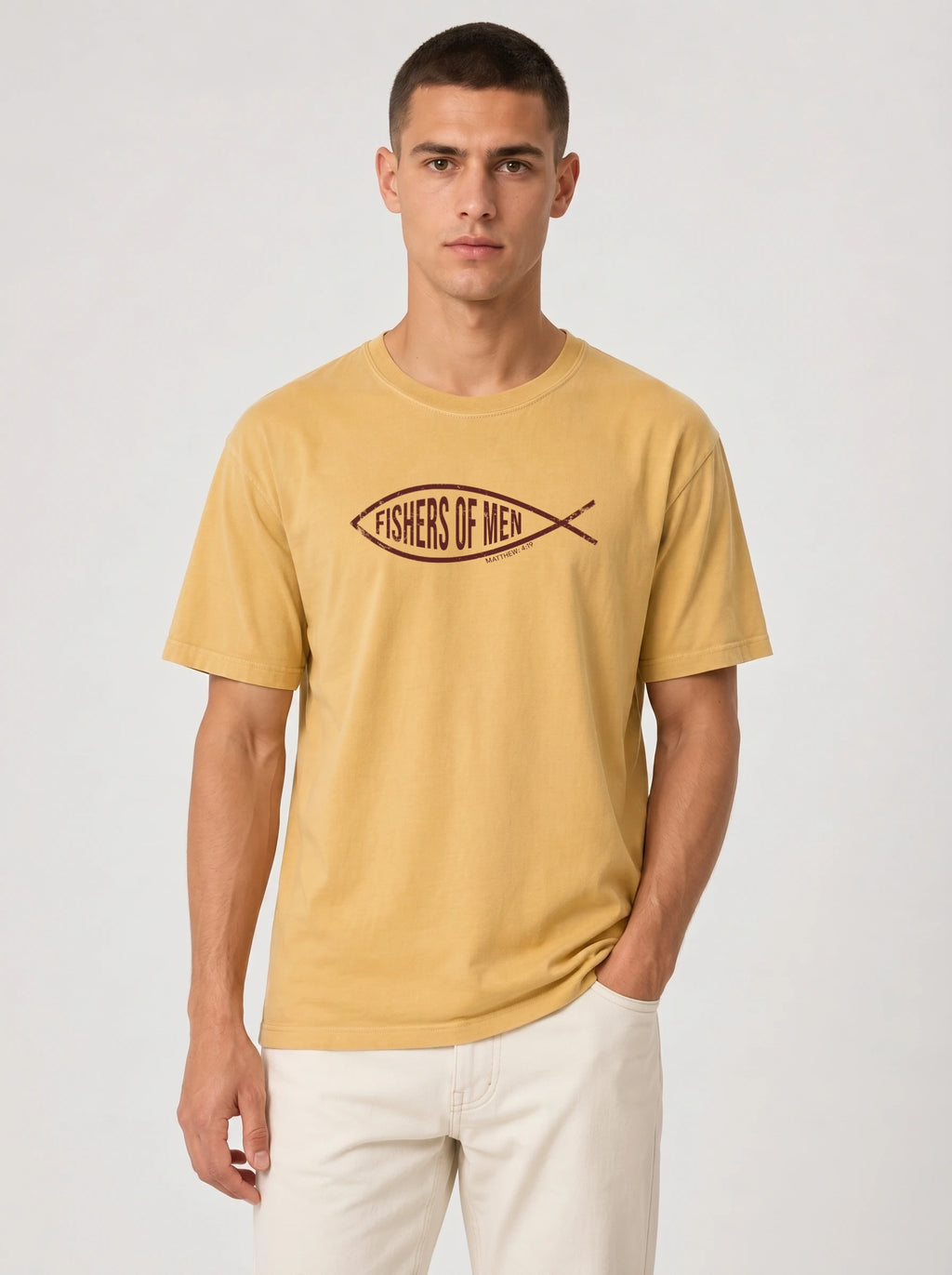 A man wearing a mustard yellow t-shirt with a dark brown 'FISHERS OF MEN' logo across the chest, paired with off-white trousers, against a light gray studio background.