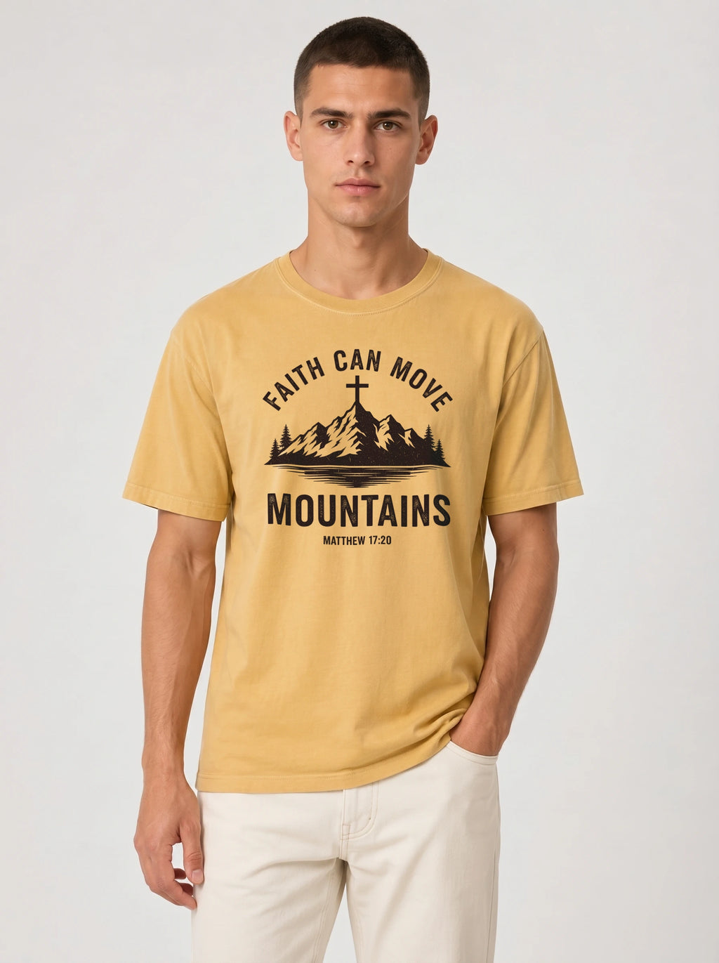 A man wearing a mustard yellow graphic t-shirt with a black mountain and cross design that reads 'FAITH CAN MOVE MOUNTAINS' and 'MATTHEW 17:20', paired with off-white pants, against a light gray studio background.