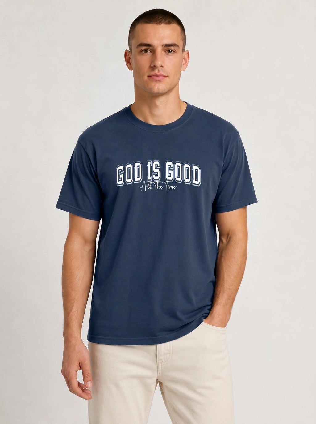 A man wearing a navy blue crewneck t-shirt with the text 'GOD IS GOOD All the time' printed in white on the chest, paired with light beige pants, against a light gray studio background.