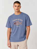 A man wearing a slate blue graphic t-shirt with a fish design and the text 'FISHERS OF MEN' and 'FISHERS OF MEN' and 'MATTHEW 4:19' on the front, paired with beige pants, against a light gray background.