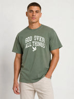 Medium front-facing shot of a male model wearing a muted sage green short-sleeve t-shirt and light beige shorts, featuring a distressed white "GOD OVER ALL THINGS" graphic and dove icon.