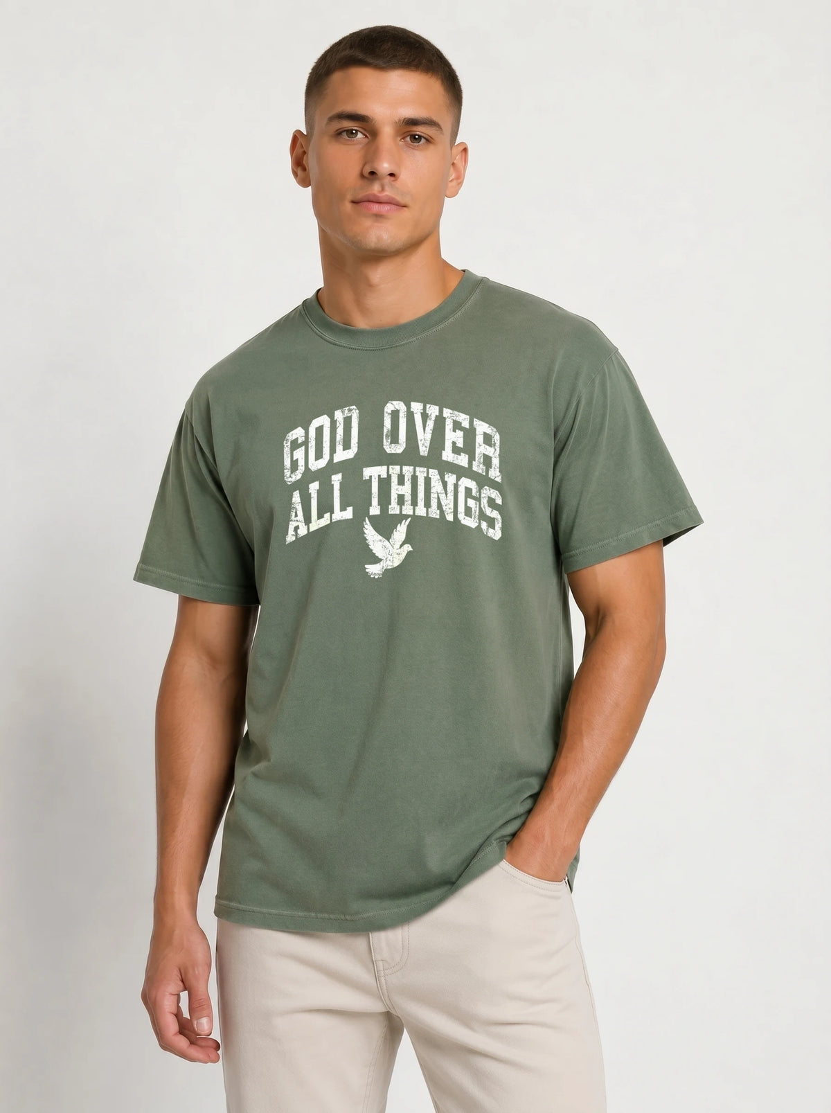 Medium front-facing shot of a male model wearing a muted sage green short-sleeve t-shirt and light beige shorts, featuring a distressed white "GOD OVER ALL THINGS" graphic and dove icon.