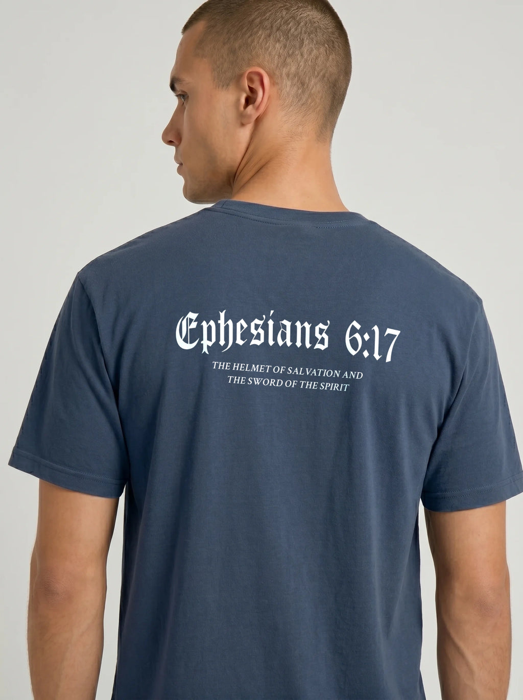 A man wearing a navy blue t-shirt, viewed from the back. The shirt features a white gothic-style print of the Bible verse Ephesians 6:17 with additional text below reading 'THE HELMET OF SALVATION AND THE SWORD OF THE SPIRIT'.