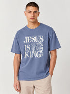 A man wearing a slate blue t-shirt with a white graphic print that reads 'JESUS IS KING' alongside a lion illustration, paired with beige pants, against a light gray studio background.