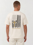 A man viewed from the back wearing a cream-colored t-shirt with a distressed black and gold American flag graphic on the back, featuring a cross and the words 'FAITH, FAMILY, FREEDOM.'.