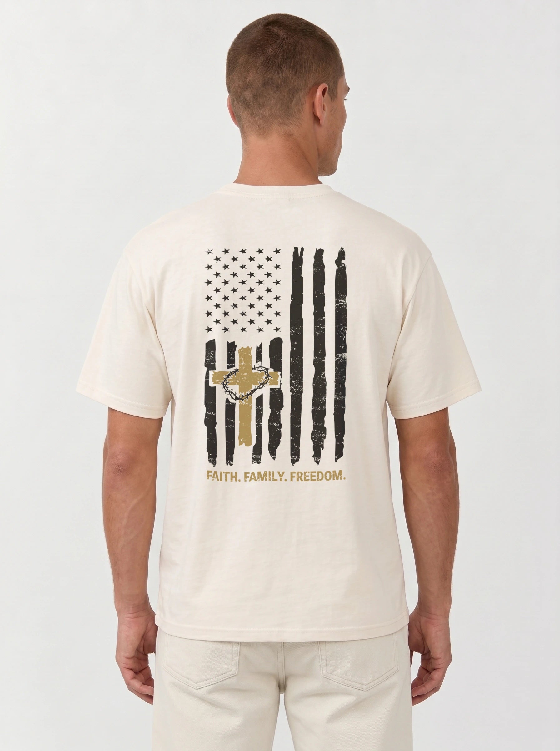 A man viewed from the back wearing a cream-colored t-shirt with a distressed black and gold American flag graphic on the back, featuring a cross and the words 'FAITH, FAMILY, FREEDOM.'.