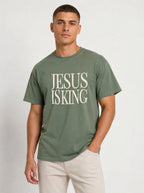 Person wearing a muted green T-shirt with distressed cream text reading “Jesus Is King.”