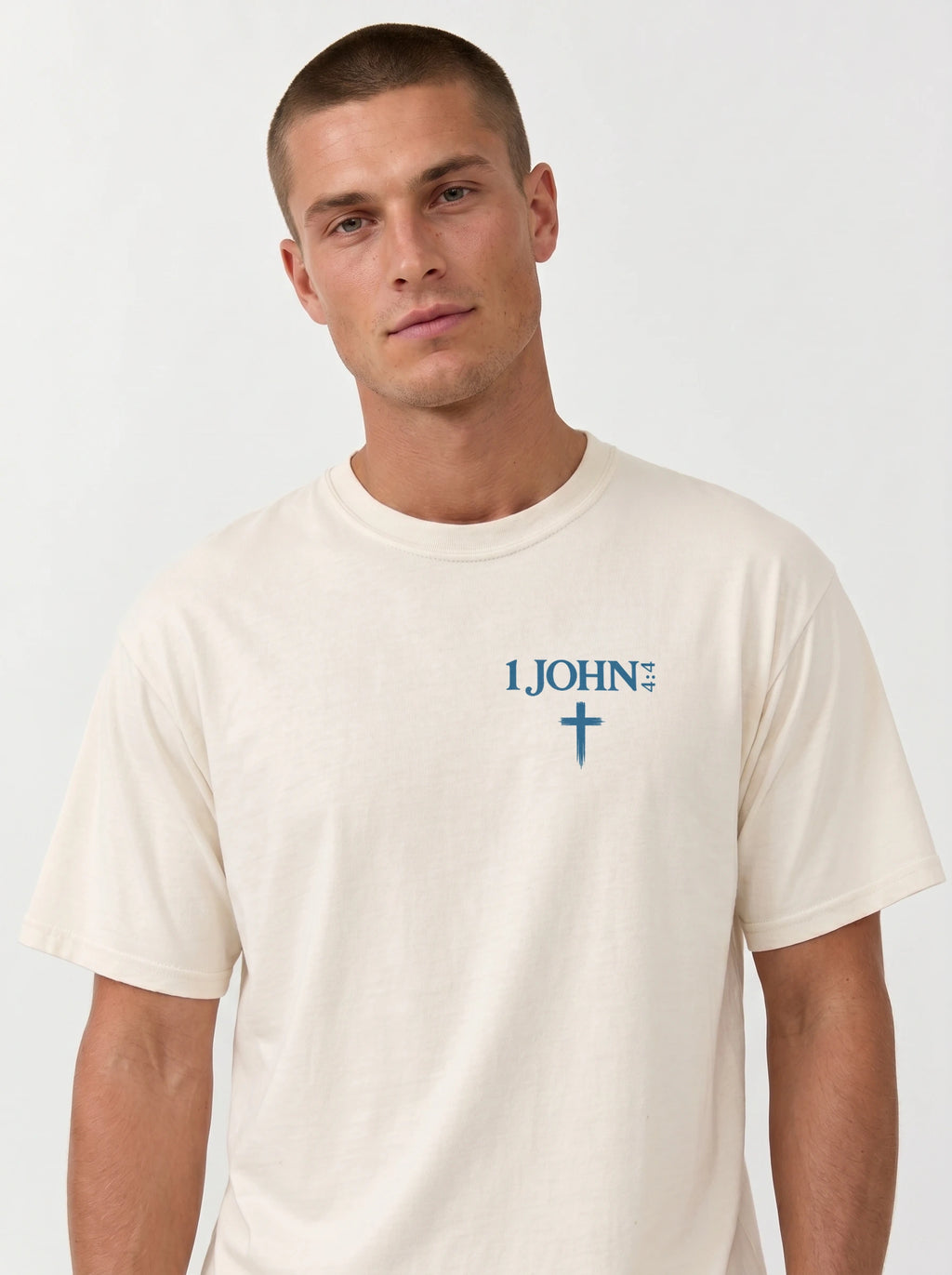 A man wearing a cream-colored garment-dyed t-shirt, viewed from the front. The shirt has a small chest print reading '1 JOHN 4:4' with a cross symbol beneath it.