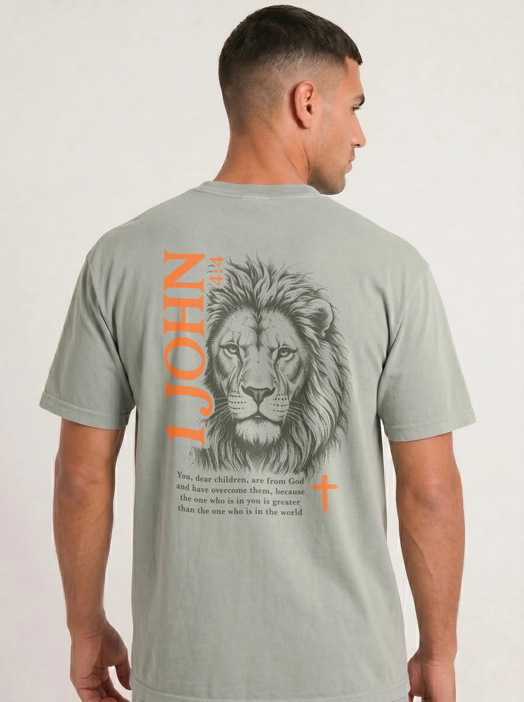 Person wearing a gray-green T-shirt with a lion illustration, orange “1 John 4:4” and cross, and the verse “You, dear children, are from God and have overcome them, because the one who is in you is greater than the one who is in the world.”