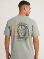 Person wearing a gray-green T-shirt with a lion illustration, orange “1 John 4:4” and cross, and the verse “You, dear children, are from God and have overcome them, because the one who is in you is greater than the one who is in the world.”