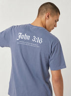 A man wearing a slate blue t-shirt, viewed from the back. The shirt features white printed text reading 'John 3:16' in a gothic font, with 'OUR GOD HAS LOVED THE WORLD THAT HE WASHES US SIN AND ONLY HOW' printed below in a smaller serif font.