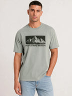 A man wearing a light sage green t-shirt with a black mountain graphic and the text 'FAITH CAN MOVE MOUNTAINS' printed on the chest, paired with light blue jeans, against a plain light gray background.