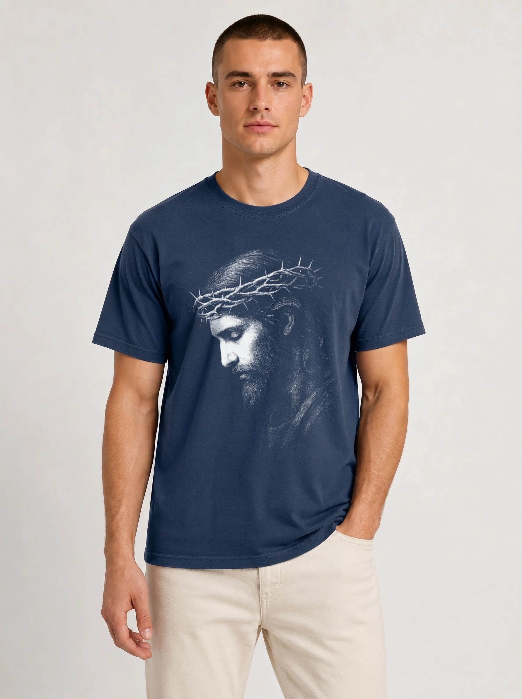 A man wearing a navy blue crewneck t-shirt with a white graphic print of Jesus' face wearing a crown of thorns, paired with beige pants, against a light gray studio background.