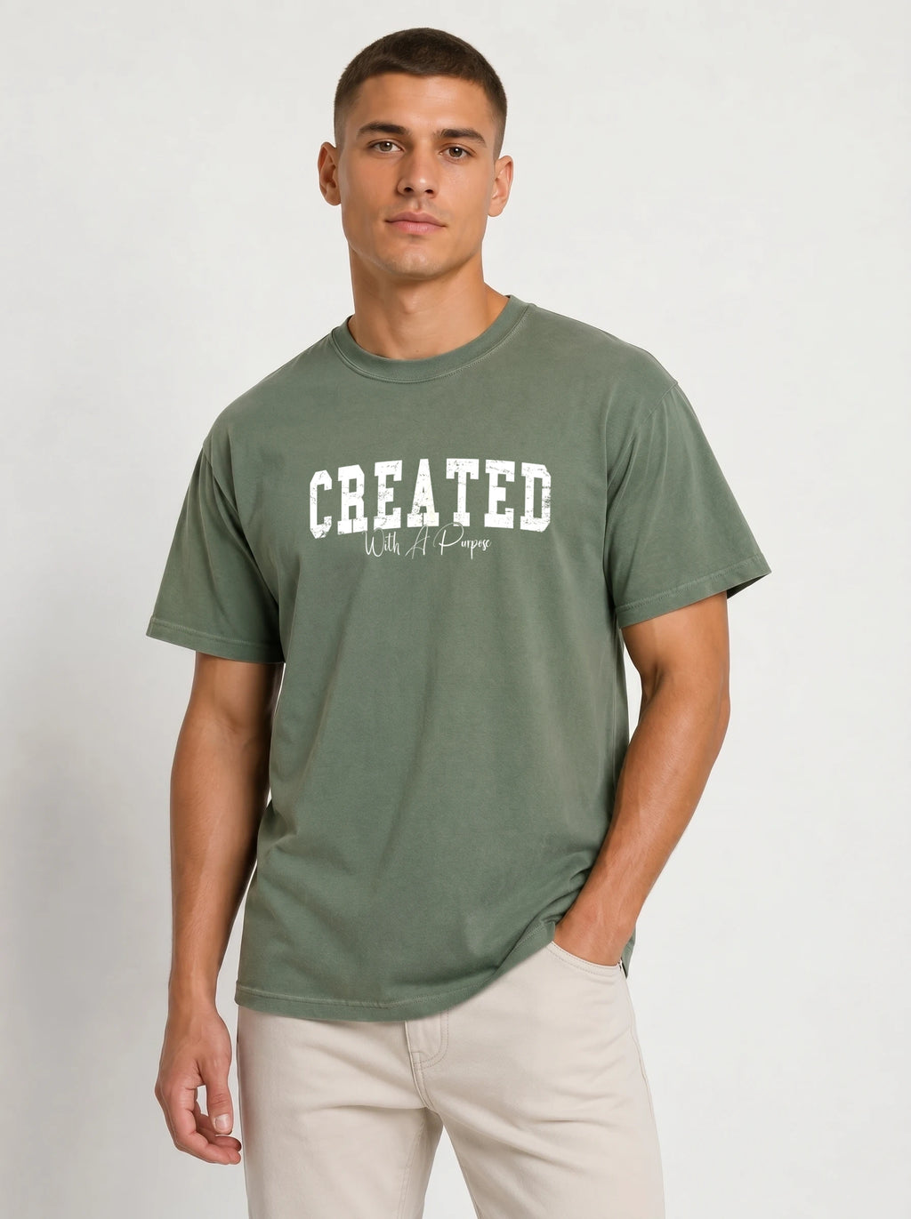 A man wearing an olive green t-shirt with 'CREATED' printed in white across the chest, paired with beige pants, against a light gray studio background.