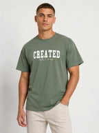 A man wearing an olive green t-shirt with 'CREATED' printed in white across the chest, paired with beige pants, against a light gray studio background.