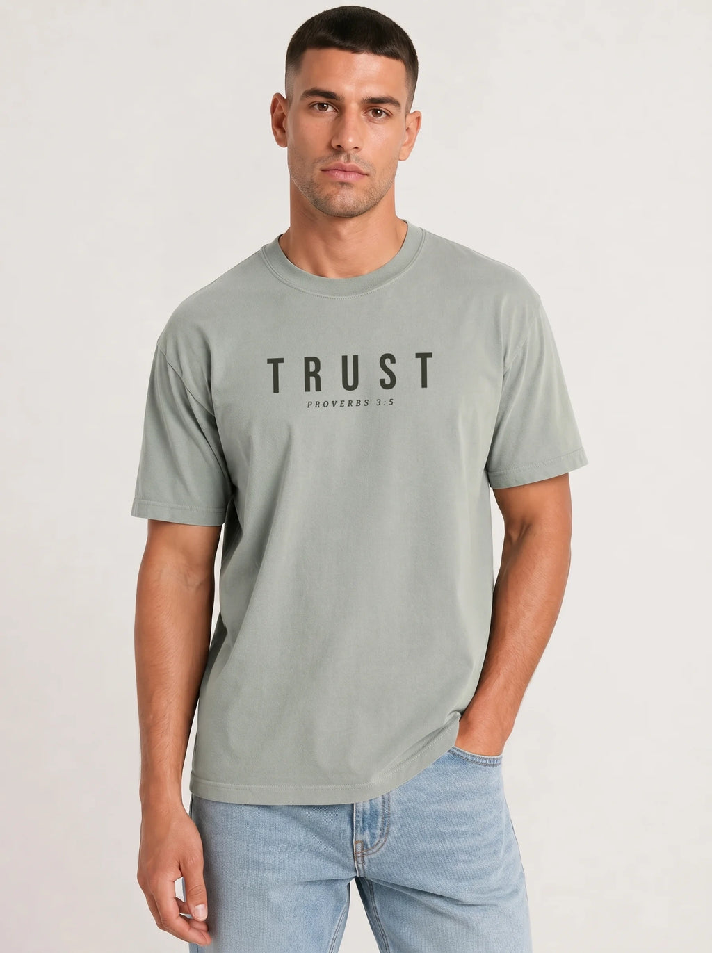 A man wearing a light sage green crewneck t-shirt with 'TRUST' printed in black across the chest, paired with light blue jeans, against a plain light gray background.