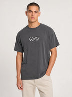 A man wearing a dark gray washed t-shirt with 'God Is Greater Than Highs and Lows' printed on the chest, paired with light beige pants, against a light gray studio background.