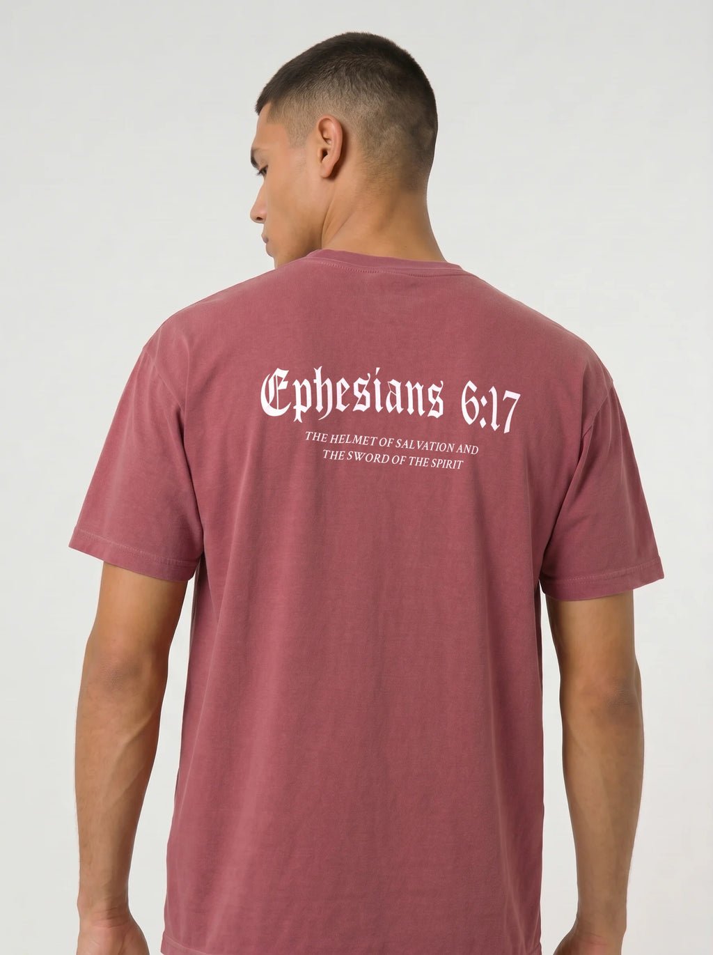 A man viewed from the back wearing a dusty rose short-sleeve t-shirt with white gothic text reading 'Ephesians 6:17' and smaller text underneath, against a light gray studio background.