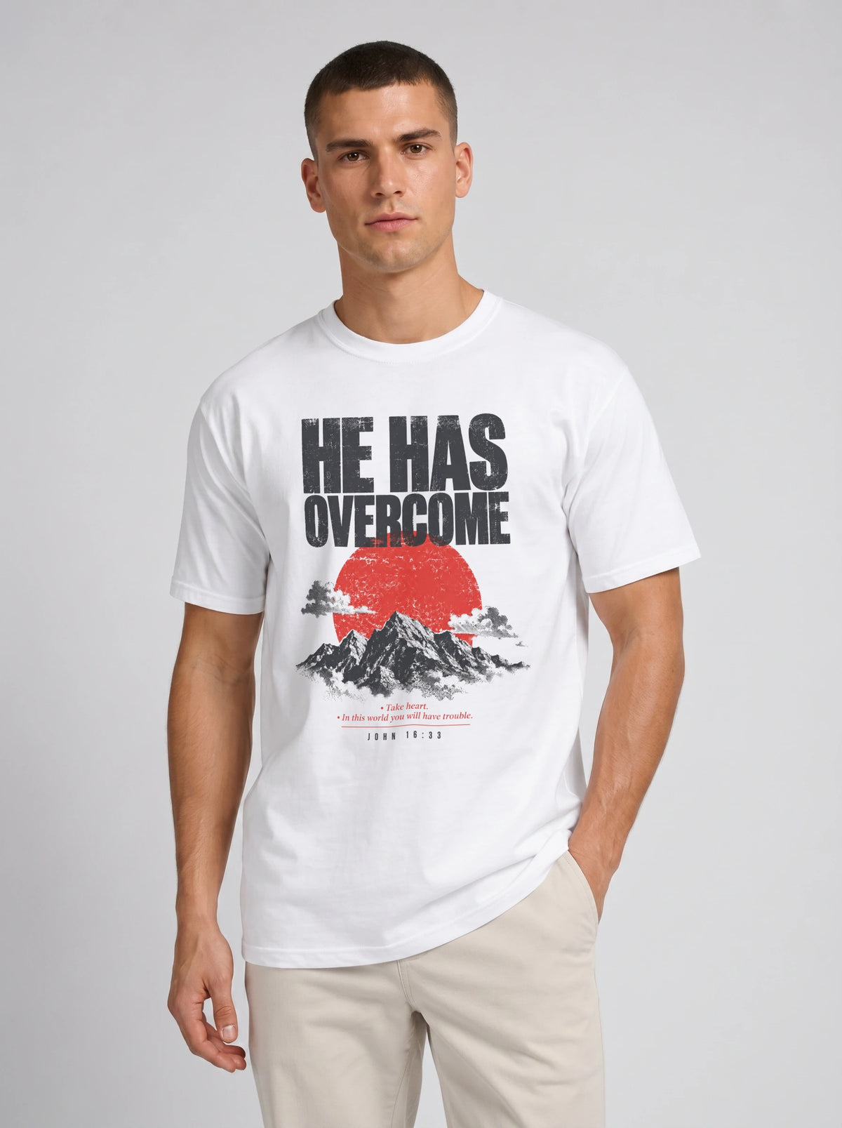 A man wearing a white graphic t-shirt with the text 'HE HAS OVERCOME' above a red sun and mountain design, paired with beige pants, against a light gray studio background.