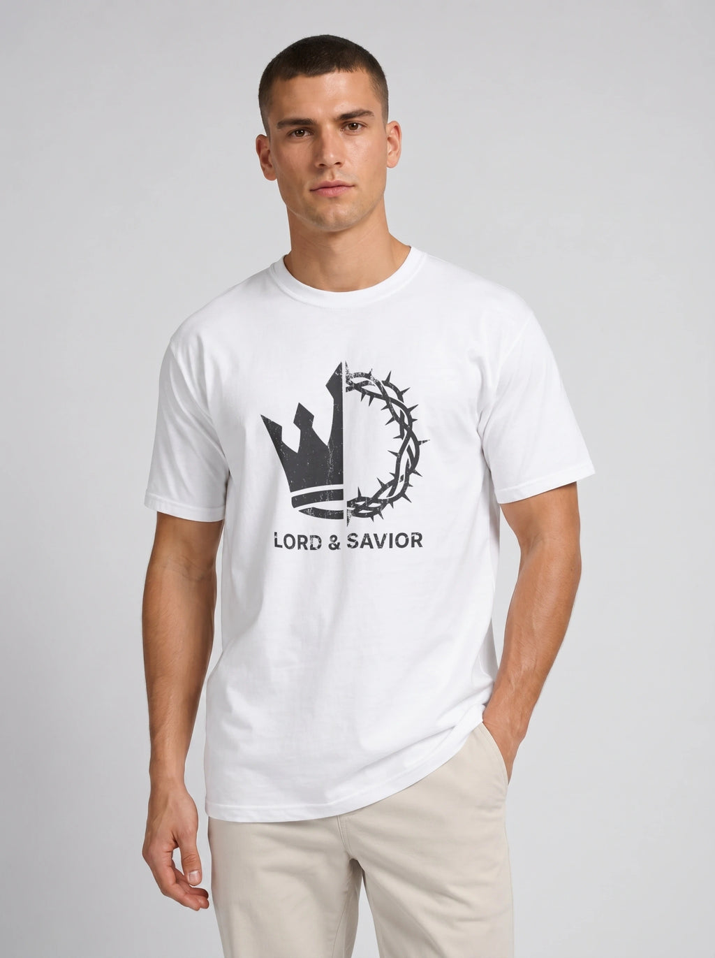 A man wearing a white crewneck t-shirt with a black graphic of a crown of thorns and the text 'LORD & SAVIOR' on the chest, paired with beige pants, against a light gray studio background.