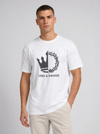 A man wearing a white crewneck t-shirt with a black graphic of a crown of thorns and the text 'LORD & SAVIOR' on the chest, paired with beige pants, against a light gray studio background.