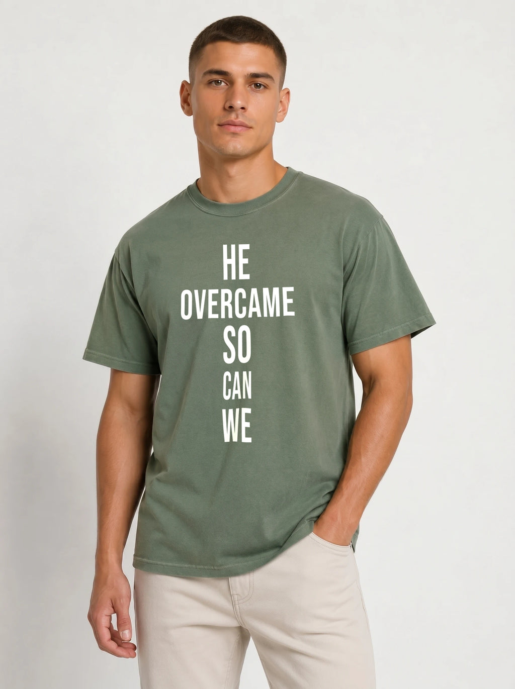 Person wearing a muted green T-shirt with large white text reading “HE OVERCAME SO CAN WE.”