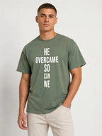 Person wearing a muted green T-shirt with large white text reading “HE OVERCAME SO CAN WE.”