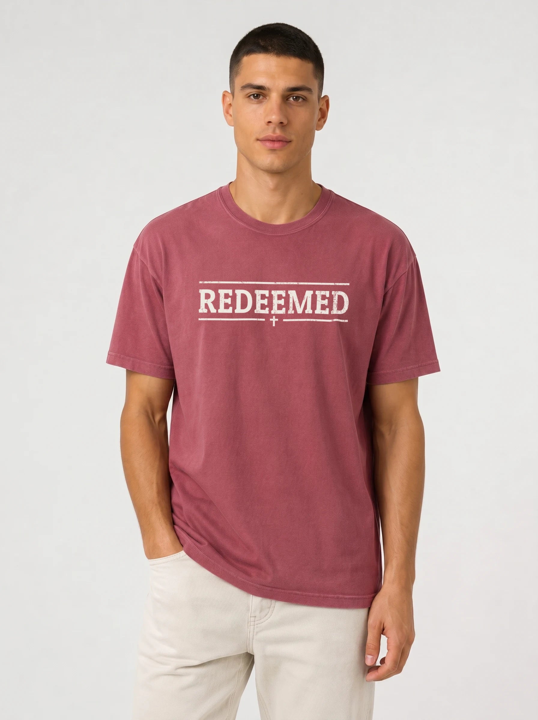 A man wearing a washed dusty rose t-shirt with 'REDEEMED' printed in distressed white block letters across the chest, flanked by two horizontal lines and a small cross below, paired with off-white pants, against a light gray studio background.