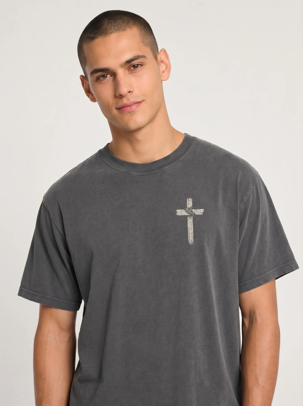 A man wearing a dark gray crewneck t-shirt with a small white cross graphic on the left chest, photographed against a light gray studio background.