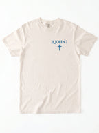 Flat lay of a cream-colored garment-dyed t-shirt, front view, laid on a white surface. It has a small chest print reading '1 JOHN 4:4' with a cross symbol beneath it.