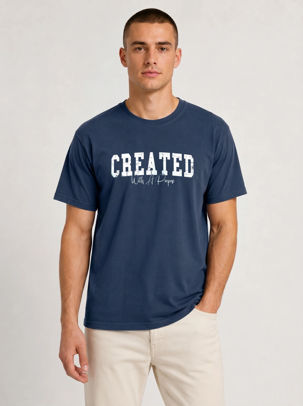 A man wearing a navy blue crewneck t-shirt with 'CREATED With A Purpose' printed in white across the chest, paired with light beige pants, against a light gray studio background.