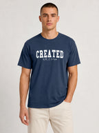A man wearing a navy blue crewneck t-shirt with 'CREATED With A Purpose' printed in white across the chest, paired with light beige pants, against a light gray studio background.