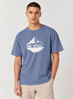 A man wearing a heathered blue crewneck t-shirt with a white mountain graphic and the text 'BE STILL' on the chest, paired with beige pants, against a light gray studio background.