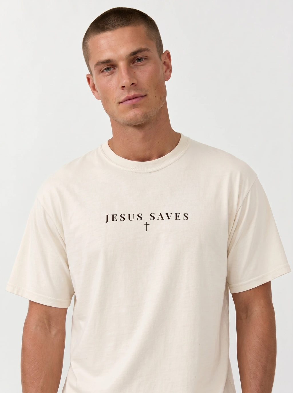 Young man modeling an off-white short-sleeve T-shirt featuring black “JESUS SAVES” text with a small cross below, posed against a plain light background.