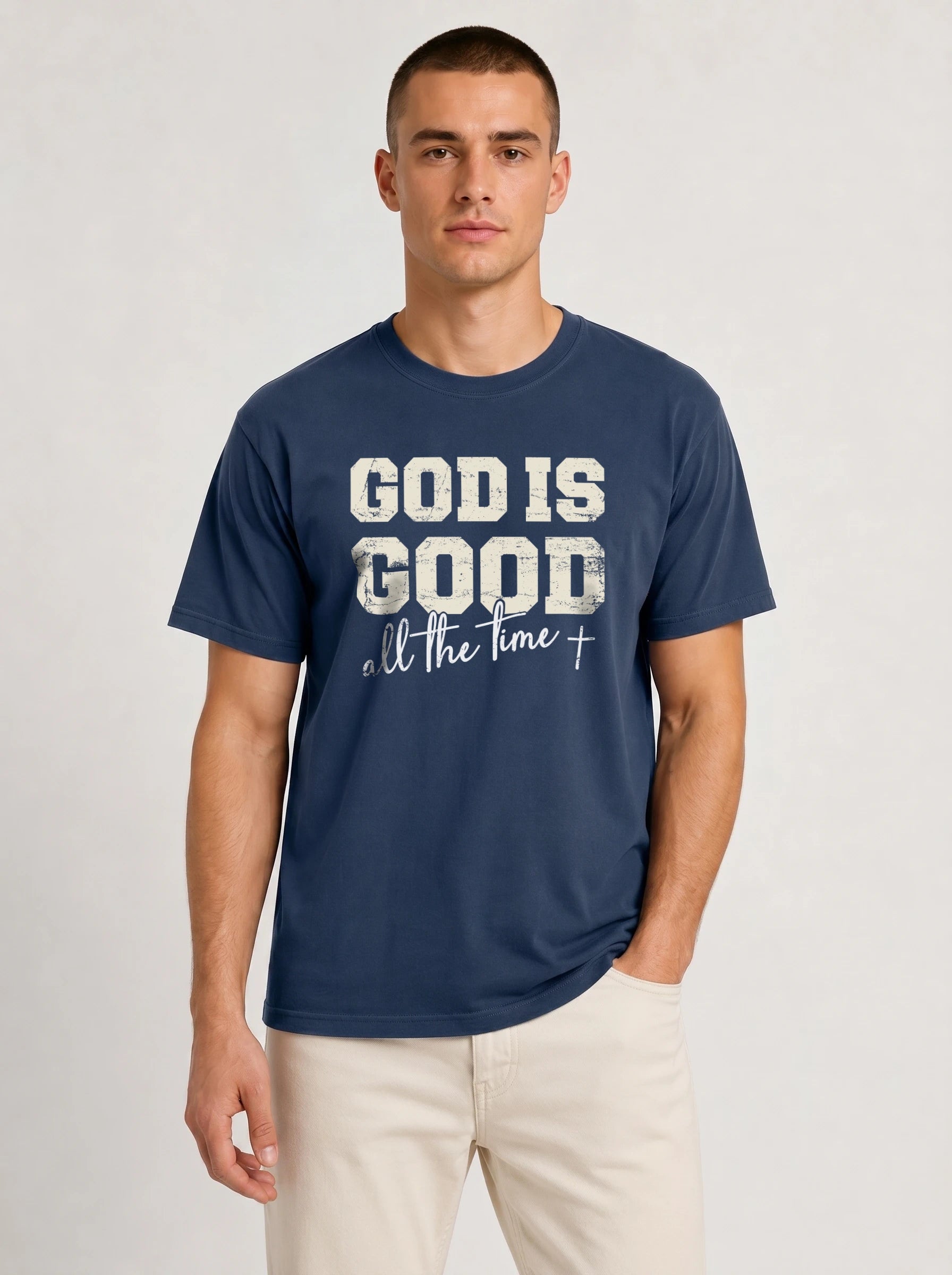 A man wearing a navy blue t-shirt with a distressed white graphic that reads 'GOD IS GOOD all the time+' paired with light beige pants, photographed against a neutral light gray background.