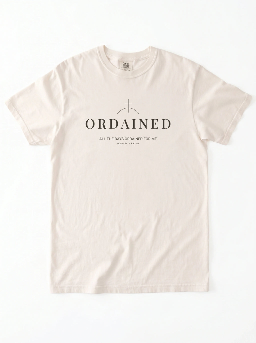 A light cream-colored short-sleeve t-shirt with a black graphic print on the chest featuring a cross, the word 'ORDAINED', and a Bible verse, displayed flat against a white background.