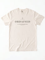 A light cream-colored short-sleeve t-shirt with a black graphic print on the chest featuring a cross, the word 'ORDAINED', and a Bible verse, displayed flat against a white background.