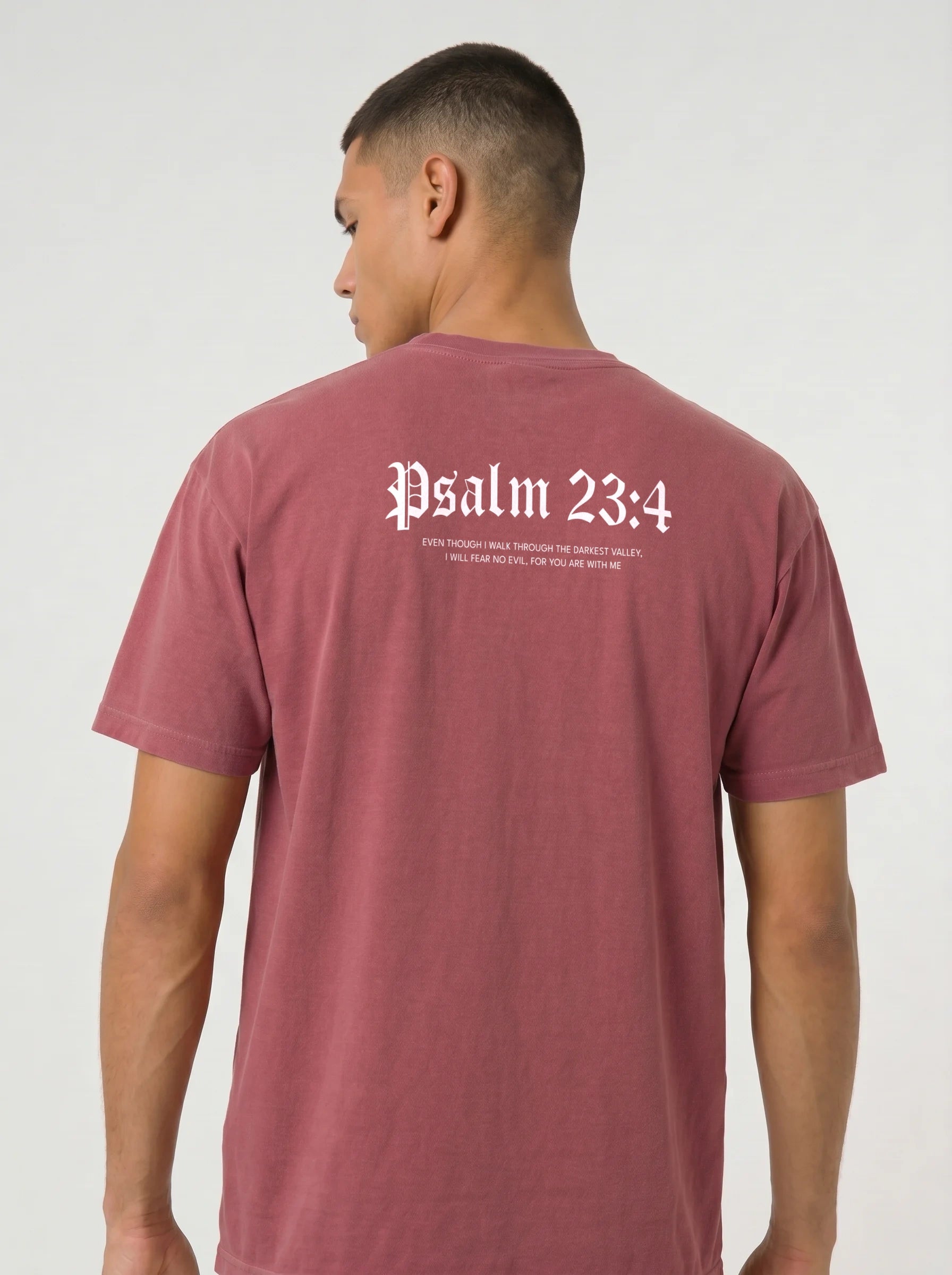 A man wearing a dusty rose short-sleeve t-shirt, viewed from the back. The shirt features the text 'Psalm 23:4' printed in a white Gothic font, with smaller white text underneath, against a plain light gray background.