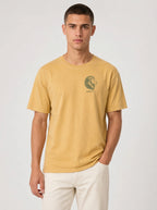 A man wearing a mustard yellow crew-neck t-shirt with a green lion graphic and 'JEREMIAH 17:7' text on the left chest, paired with off-white trousers, against a light gray studio background.