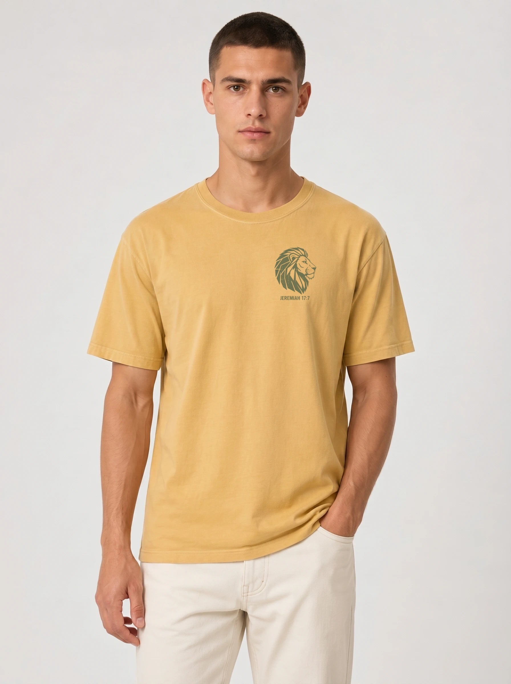 A man wearing a mustard yellow crew-neck t-shirt with a green lion graphic and 'JEREMIAH 17:7' text on the left chest, paired with off-white trousers, against a light gray studio background.