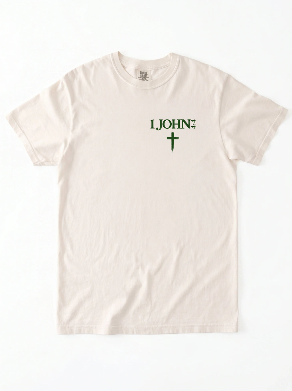 Cream T-shirt laid flat with green “1 John 4:4” text and a cross printed on the chest.