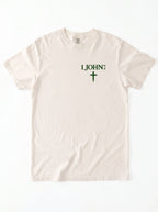 Cream T-shirt laid flat with green “1 John 4:4” text and a cross printed on the chest.
