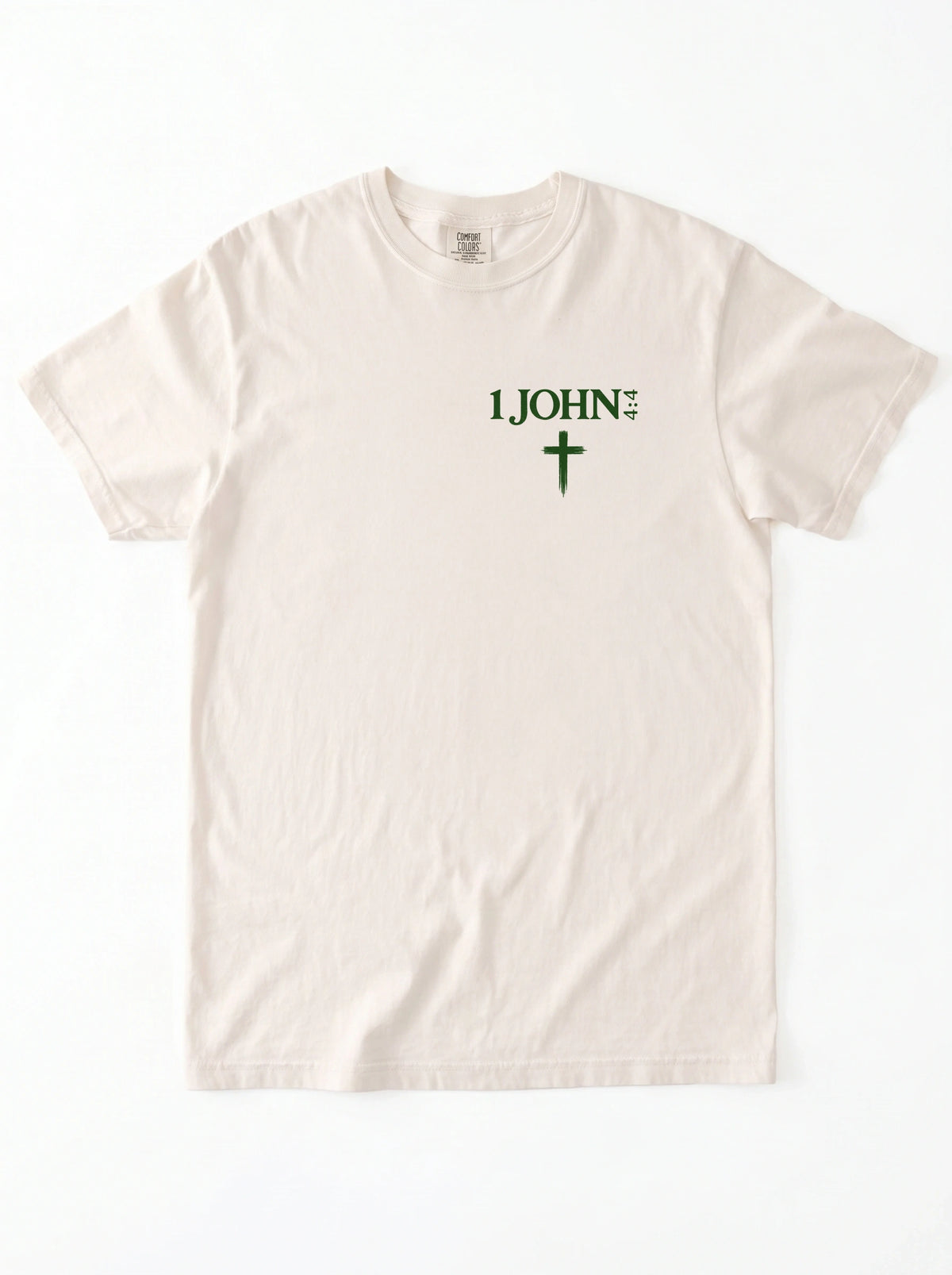 Cream T-shirt laid flat with green “1 John 4:4” text and a cross printed on the chest.