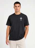 A man wearing a black short-sleeve crewneck t-shirt with a small white cross printed on the left chest, paired with light beige pants, against a plain white studio background.