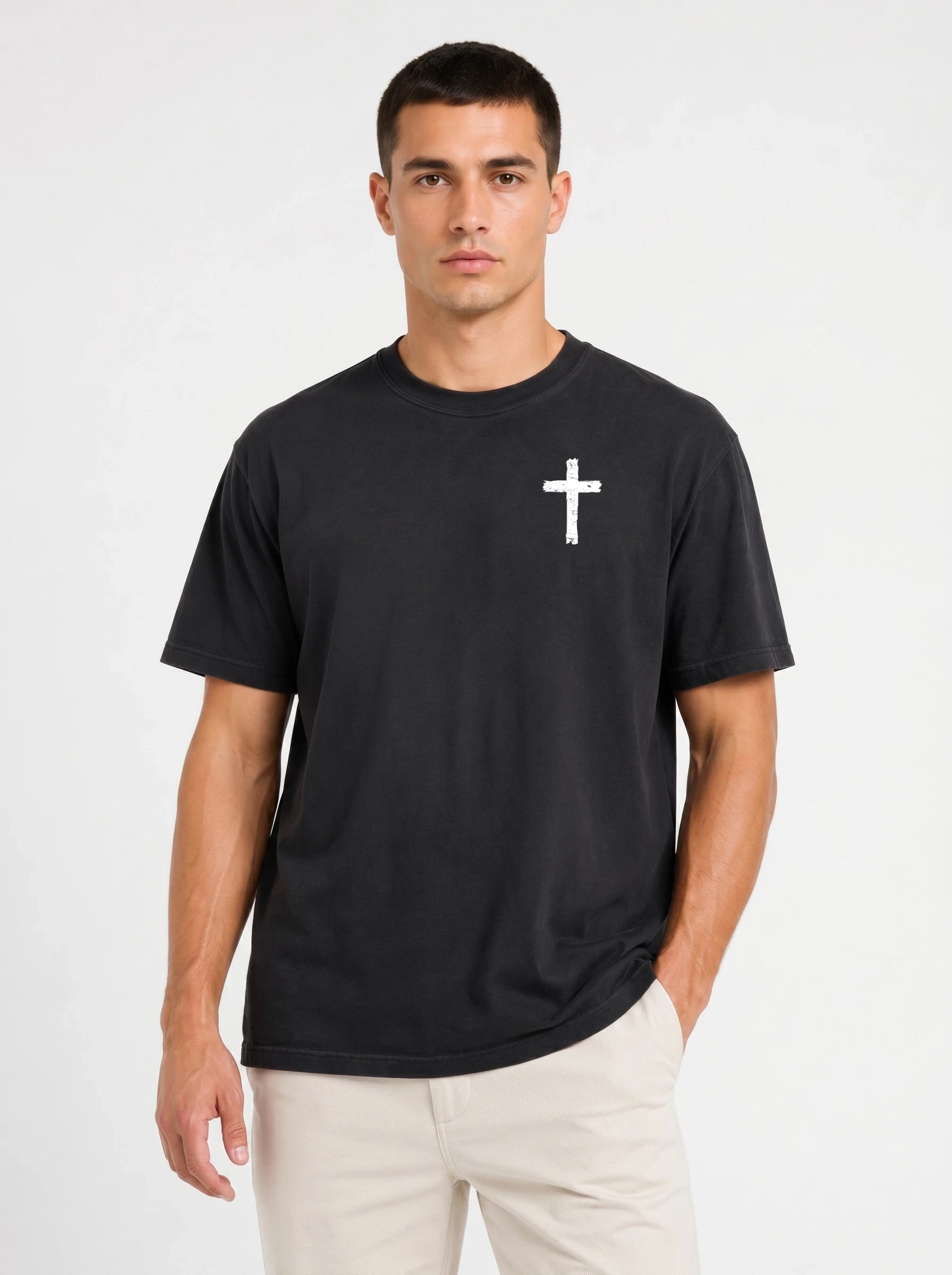 A man wearing a black short-sleeve crewneck t-shirt with a small white cross printed on the left chest, paired with light beige pants, against a plain white studio background.