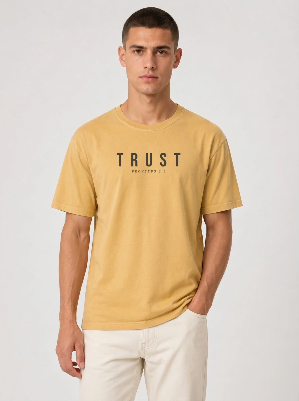 A man wearing a mustard yellow crewneck t-shirt with 'TRUST' printed in black across the chest, paired with off-white pants, against a light gray studio background.